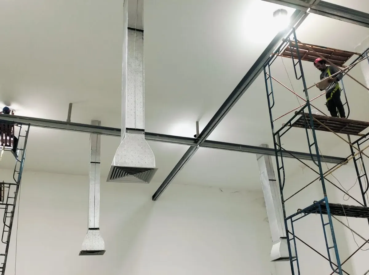 Teknisi di atas scaffolding memasang ducting galvanis di ruang produksi berdinding putih